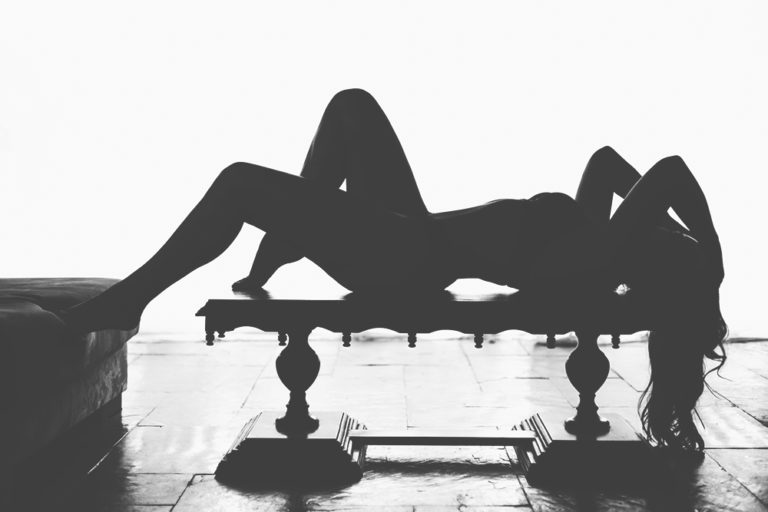 Foto sensual de mulher deitada em cima de uma mesa em imagem preto e branco de ensaio boudoir em contraluz