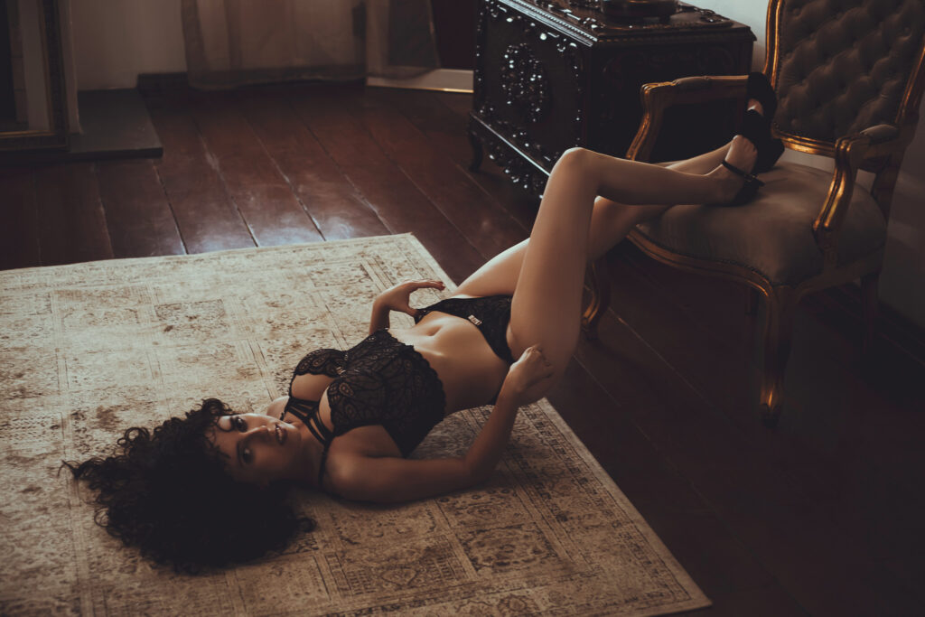 Foto sensual de mulher de cabelos cacheado e vestindo lingerie preta deitada no chão em sala de estúdio fotográfico para ensaio boudoir